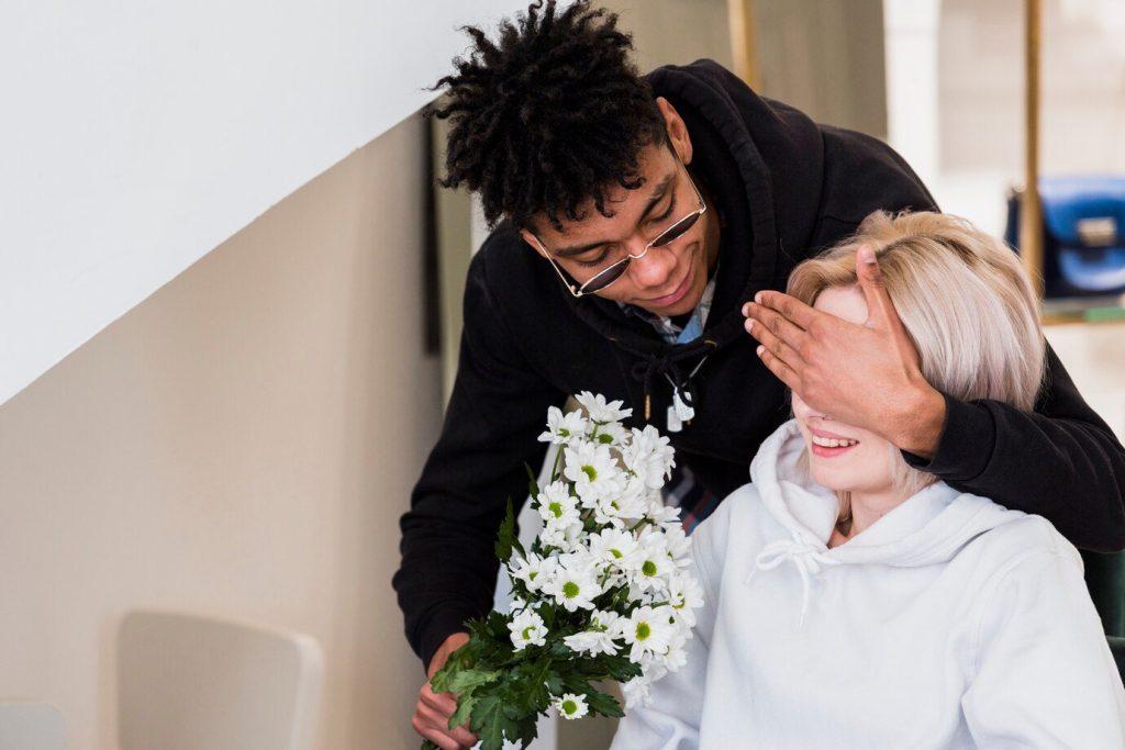 young man closing her girlfriend's eye holding flower bouquet in hand