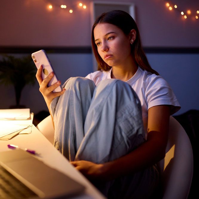 Worried Teenage Girl Sitting At Desk In Bedroom At Home Looking At Mobile Phone At Night