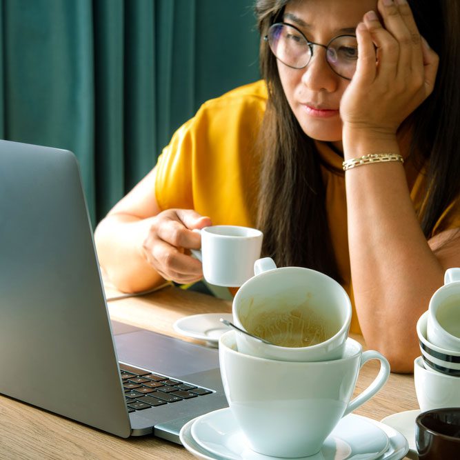 Asian woman holding a cup of coffee sitting working frustrated exhausted looking at laptop screen.