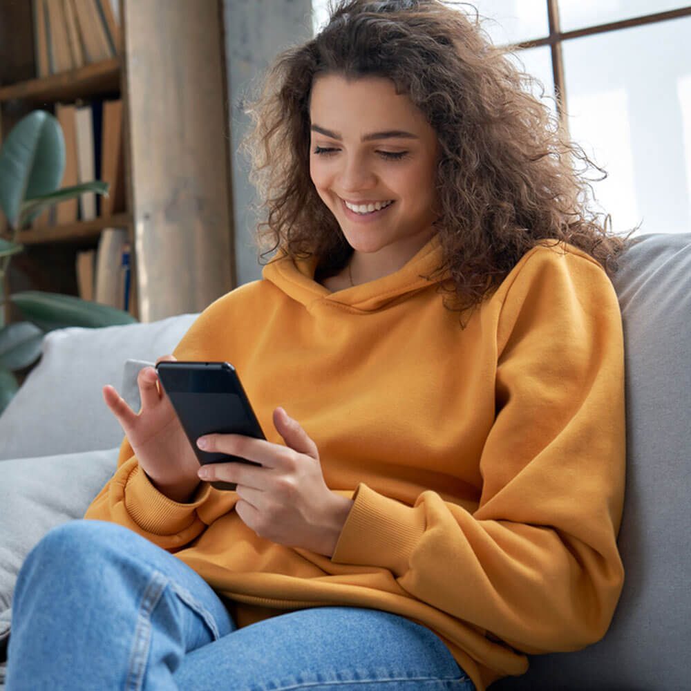 Person in a yellow hoodie using a smartphone on a couch, finding a therapist in NYC.