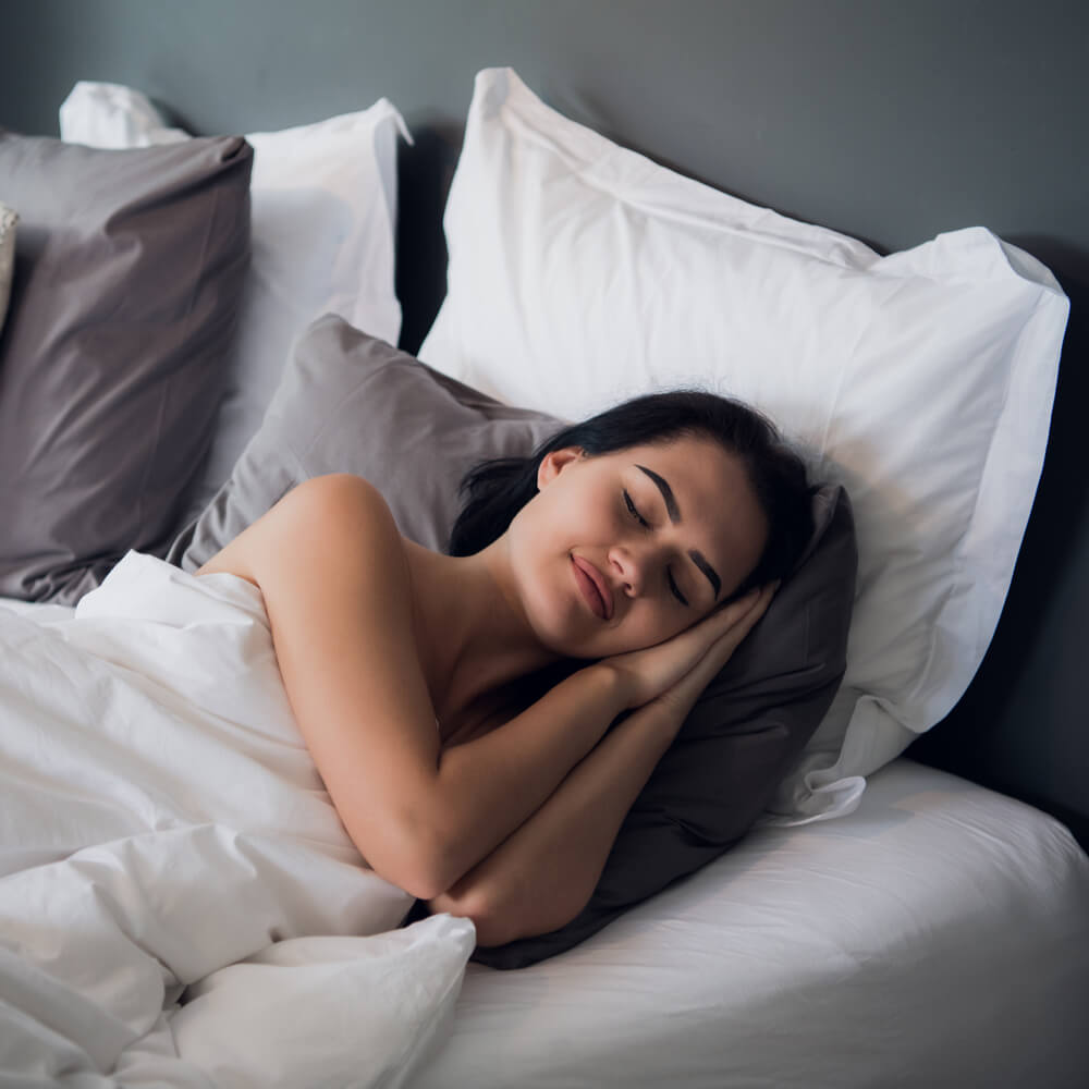 Person curled up in bed with white and grey pillows, focusing on better sleep through CBT in NYC.