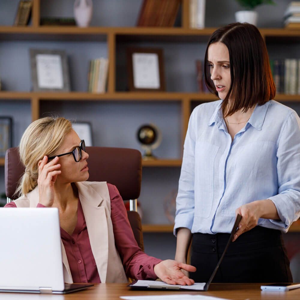 Two women at a desk, one with hand on temple, highlighting signs of a toxic boss in NYC.