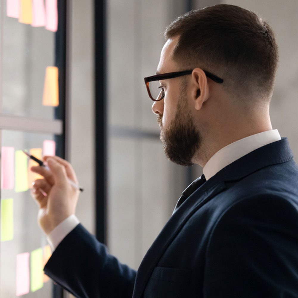 Man in a suit with a pen examining sticky notes on a glass wall, setting goals for employees in NYC.