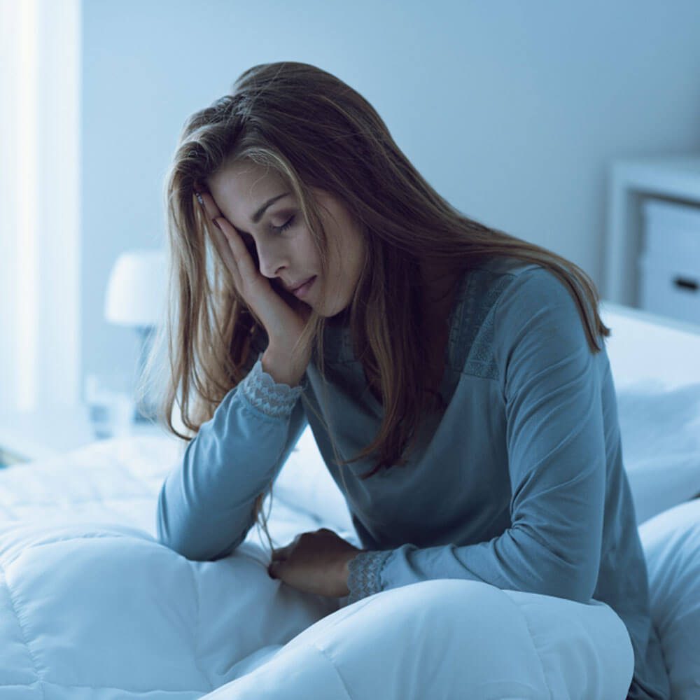 Person with head in hand on a bed in a dim room, dealing with insomnia during their period in NYC.