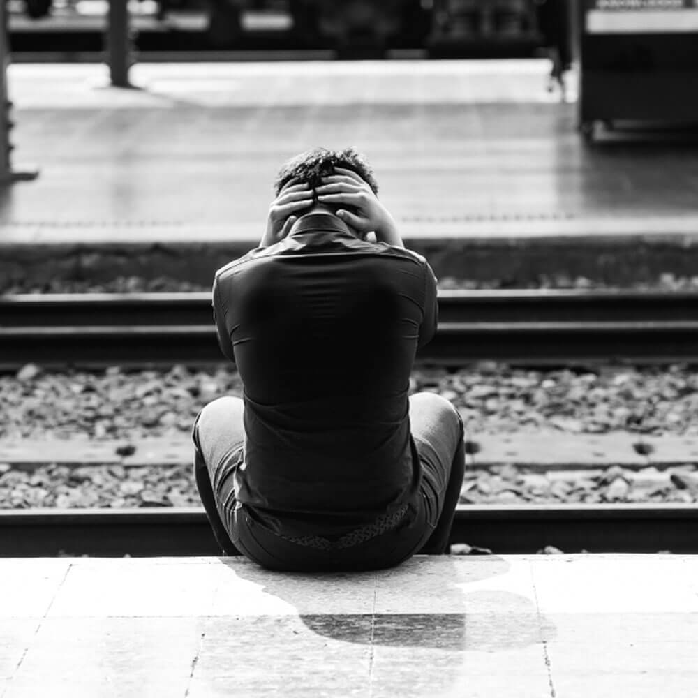 Person sitting by train tracks with hands on head, reflecting on suicidal warning signs in NYC.