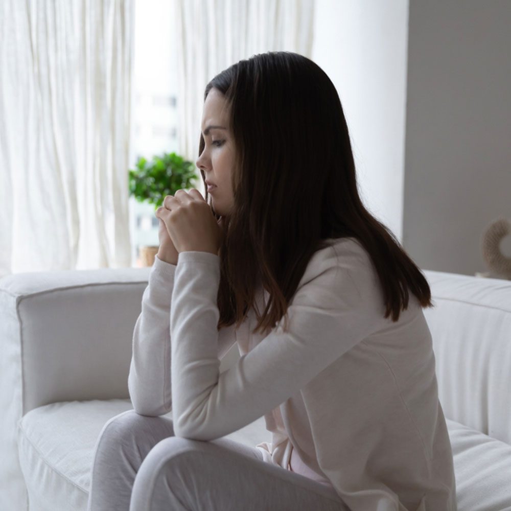 Woman in white sitting on a couch, clasping her hands, reflecting on being cheated on in NYC.
