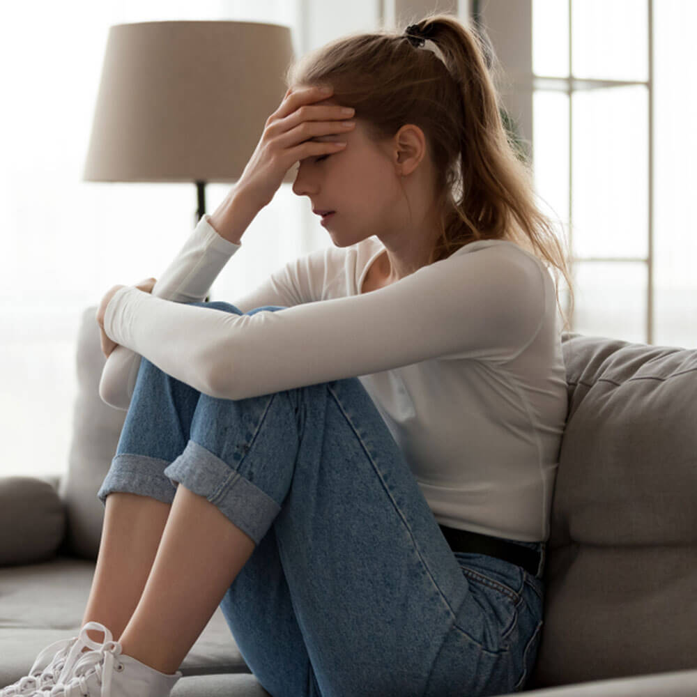 Woman sitting on couch with knees drawn up, signs of repressed childhood trauma in adults in NYC.