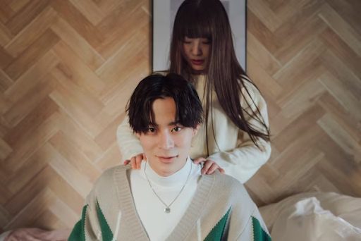 A person sitting on a bed while another stands behind, hands on their shoulders.