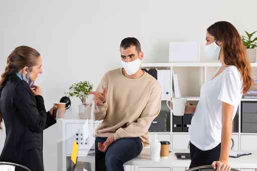 Three people in an office, one seated on a desk, while two standing with coffee mugs.