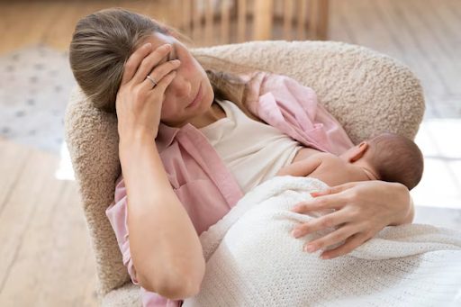 A tired mother holding her forehead while breastfeeding a baby in a cozy chair.