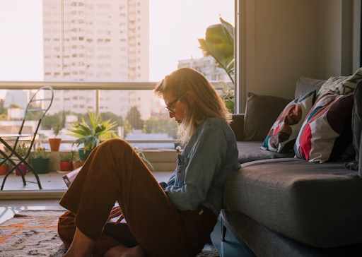 Person sitting on the floor by a sofa near a balcony with plants, during sunset.