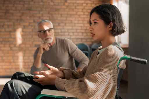 Two individuals seated indoors, one in a wheelchair, in a conversation.