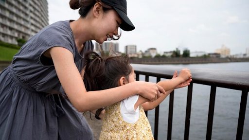 Adult and child leaning on a railing by a river, with urban buildings.