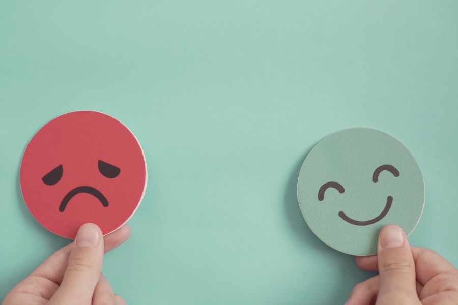 Two hands holding up a sad red face and a happy green face against a teal background.