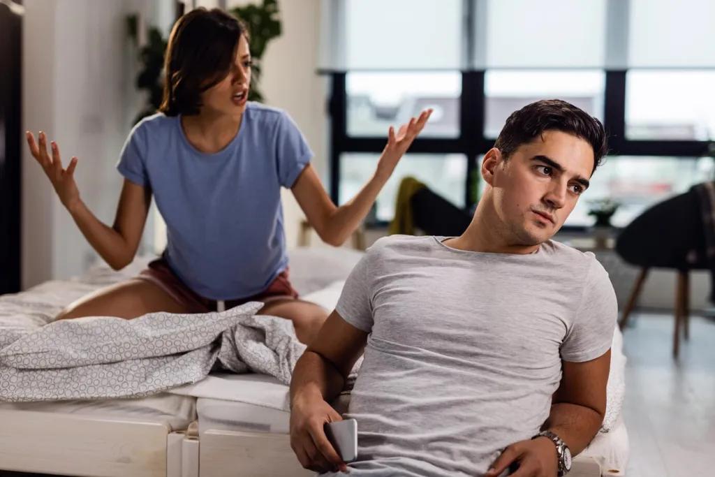 Young man sitting by the bed and ignoring his girlfriend who is arguing with him