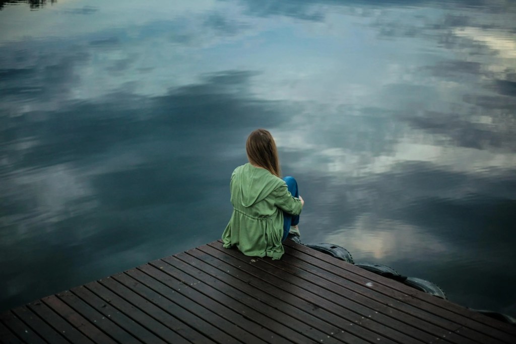 A young woman sits on a wooden dock, gazing contemplatively at the calm.