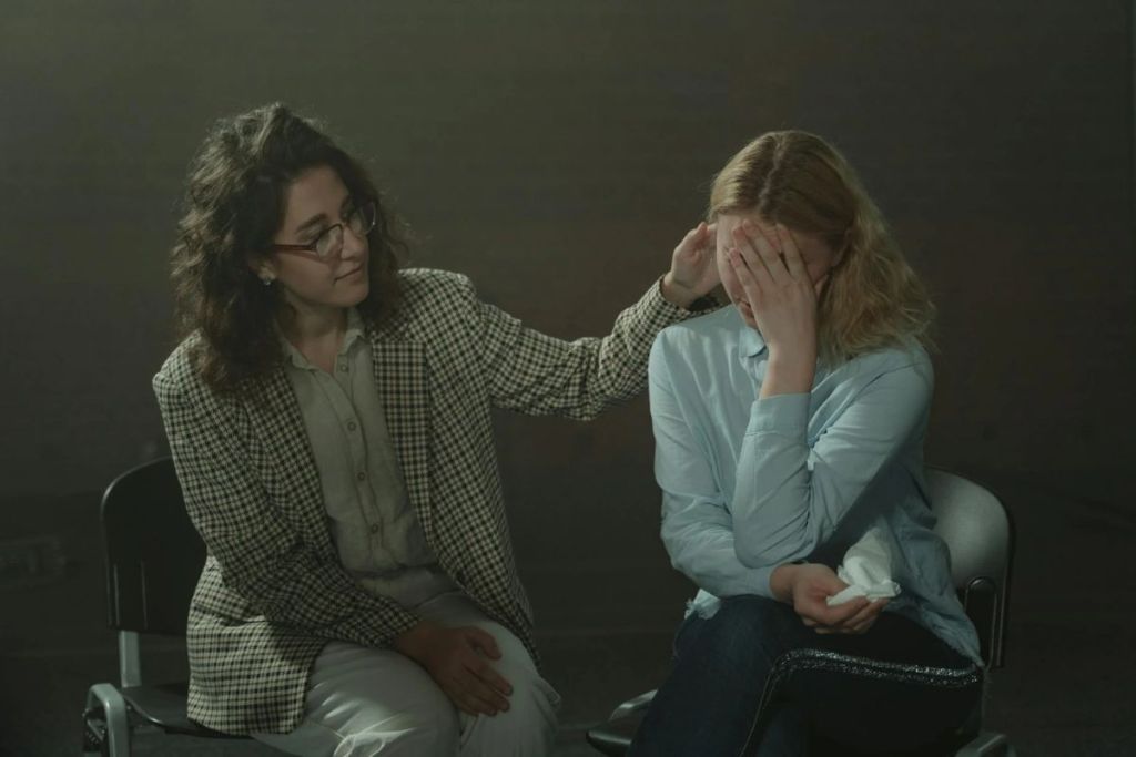 A woman comforts another who is visibly upset, seated in a dimly lit room.