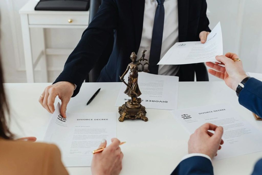 Two individuals exchange divorce documents at a table, with a statue of Lady Justice in the center.